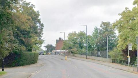 Trees line the road on Bramhall Lane with some houses seen in the background.