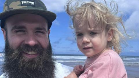 A man with a hat and a large beard takes a selfie at the beach, with the sea in the background. He is smiling while holding a young girl, who has blonde hair and a pink top.