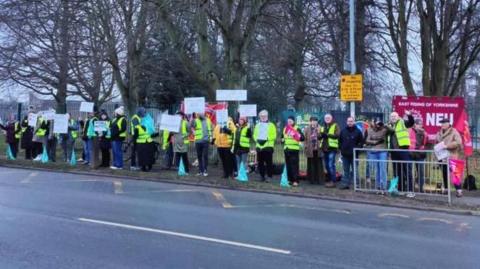 A long line of people wearing hi-vis jackets standing on the opposite side of a road on a picket line. They are standing in front of banners and many are holding white signs.