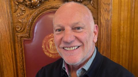 Paul Mackings smiling into the camera while sitting on a dark wood and red ornate chair. He is mostly bald with bits of white hair around the side of his head and grey stubble around his jaw. He is wearing a dark jumper with a black and white checked collar poking out underneath.