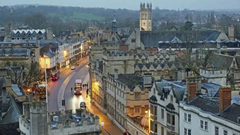 An elevated view of the historic Oxford city centre. The city's medieval buildings are lit by the low evening light and a bus can be seen driving down the city's High Street. South Park can be seen in the background