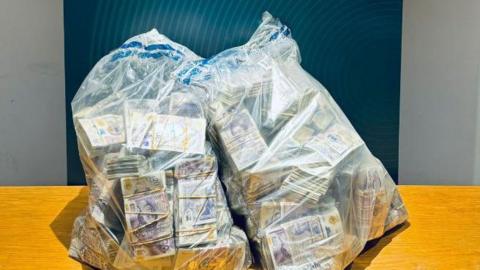 Two transparent bags sit on a wooden table - inside them bundles of £20 notes can be seen