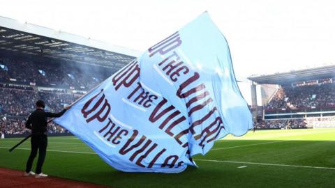 A blue Aston Villa flag is waved inside the stadium before a match. It says Up the Villa on it in maroon writing. Fans can be seen in grandstands around the pitch.
