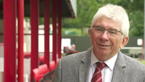 A man with white hair and glasses, a grey blazer, white shirt and a red and white striped tie, stands by a red dugout. 