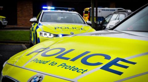 Library image of two police cars. They have yellow liveries. The vehicle in the background has its blue lights flashing.