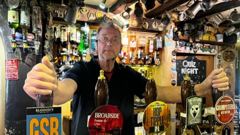 A man stood behind a pub bar. He has his hands rested on the pumps in front of him.
