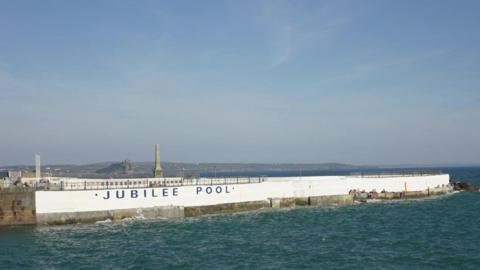 The Jubilee Pool from the outside. There is a white wall surrounding the pool and there is sea around it.