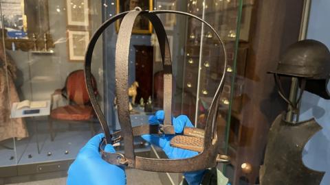 A head-shaped iron device with a plate to hold the tongue. It is being held by a person with blue plastic gloves. 