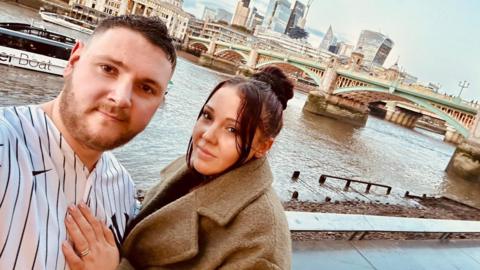 Joshua Wooldridge with his wife. They are in London and the Thames is in the background as well as an iconic bridge. There are some skyscrapers in the background. 