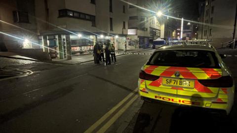 A police car parked on the pavement in the Cowgate in Edinburgh. There is a double yellow line on the road. A police officer stands in front of the fence talking to several people in dark clothing.