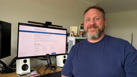 Sean Brannon with short dark hair seated at a home office desk beside a large computer monitor displaying the Buckinghamshire Plan Explorer website. The workspace includes a webcam mounted above the screen, a microphone on the desk, white speakers, a keyboard, and shelving with books and ornaments in the background. The room is brightly lit with natural light.