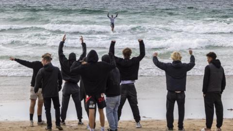 Several highly-animated people on a beach look out at a surfer. Most of them are waving their arms. The surfer has his arms up in celebration.