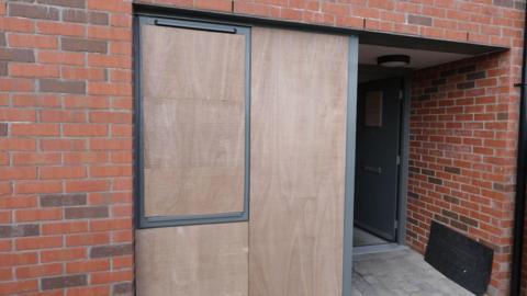 A close up of the entrance to a house with its windows boarded up in north Belfast following sectarian attacks on 21 May.   The front door of the property is open and the building is made of new red brick. 