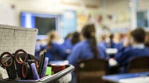 A stationary holder with various items sticking out of it with students wearing blue jumpers in a classroom looking ahead at ta screen in the background.