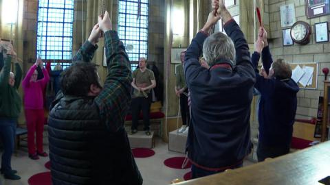 A group of people standing in a circle pulling on ropes which are attached to bells above a church.
