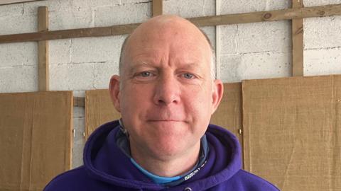A man wearing a purple hoodie stands against a white wall looking straight at the camera.