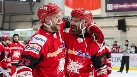 Two ice hockey players in red tops with black text on the back embrace and smile as they stand on an ice rink. Other players are visible in the background