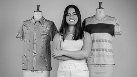 Holly poses in a black and white picture alongside some fashion designs on mannequins. The one on the left is a buttoned up collared shirt with a leaf motif. The right one is a v-neck women's shirt with square patterns. Holly is smiling, has long dark hair, and wears a white vest and white jeans.