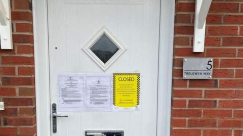 A white front door with three A4 notices stuck it, two white, one yellow. A sign on the wall next to the door says "5 Trelowen Way". The door has a diamond-shaped window.