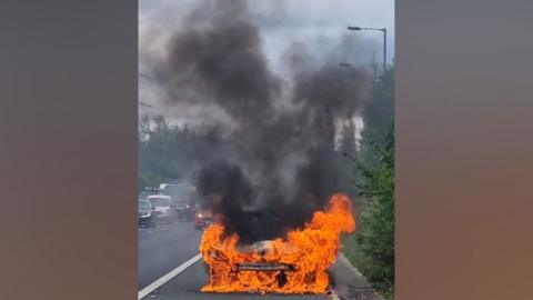 Blazing car engulfed in bright orange flames and thick black smoke on a motorway hard shoulder.