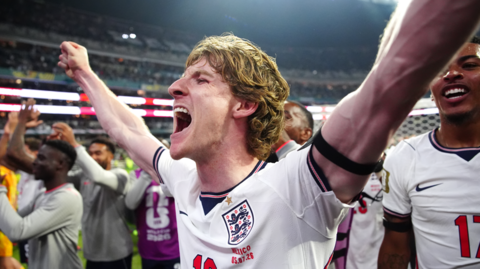 A young man with thick, curly blonde hair, wearing a white football kit with the England three lions badge on the left, stands in a football stadium cheering with his arms held aloft.