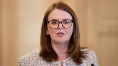 Caoimhe Archibald has shoulder length ginger hair and large dark framed glasses. She is wearing a beige coloured blazer.