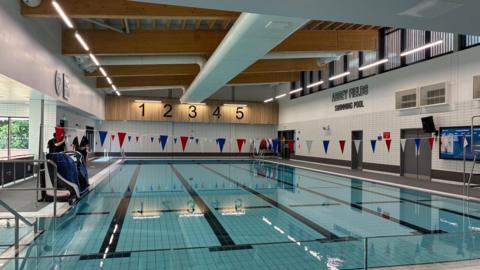 A swimming pool with five lanes and the numbers of the lanes on the far wall. The words Abbey Fields swimming pool is on the wall to the right and there is red, white and blue bunting hanging over the pool. On the left of the pool two men dressed in black stand behind the hoist to allow disabled access.