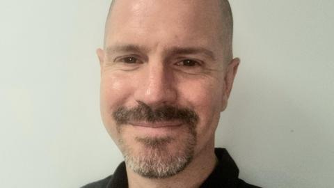Ross Ayres has brown eyes, a shaven head, moustache and cropped goatee beard. He is wearing a black top.