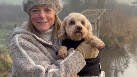 A woman, Louise Lear, holds a dog. They pose in front of a bridge over water and bushes. She wears a furry grey hat and woolly jacket. The dog wears a black body warmer