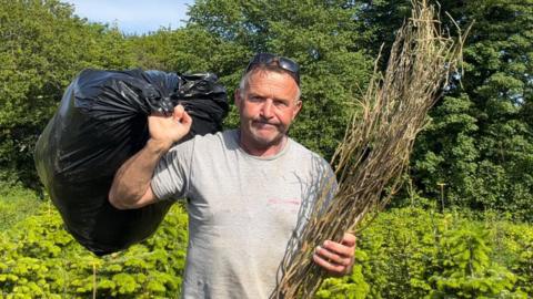 Rob Morgan is holding a black bin bag over his left shoulder and a garden waste in his right hand. He is wearning a grey t-shirt and has black sunglasses on top of his head. He is not smiling, with a fed up expression. 