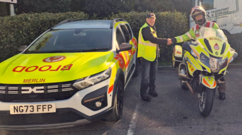 A yellow and white car with BLOOD written on it and the word Merlin. A man wearing a yellow high vis vest and dark trousers shaking hands with a man wearing a helmet leaning over a yellow blood motorbike