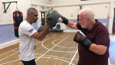 Geoff Perry, 75, is on the right with boxing gloves. He is wearing a maroon t-shirt and has short white hair, a beard and a moustache. He is punching with his right hand a pad held by coach, Paul Gill. They are in a room with a wooden floor which has white lines painted on it for various sports. Punch bags are hanging on the walls. Paul Gill is in a white t-shirt. He has cropped grey hair and a beard and moustache.