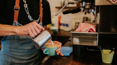 A barista making a coffee. The barista is pouring milk into the coffee mug. 