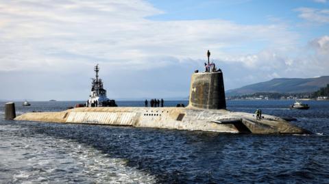 A Vanguard class submarine, which is very dirty and stained looking, is on the surface with a Scottich backdrop. There are personnel on deck and tugs beside it on a sunny day with calm seas.