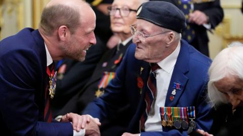 An elderly man in a blue suit, wearing military medals, sits in a wheelchair as he is shakes hands with Prince William. Other people can be seen sat beside him.