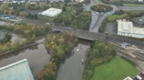 A aerial view of a flooded road with a road running over it and green fields and trees on either side