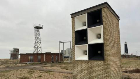 A brick rectangular-type building with six black and white foghorns inset is in the foreground. There is a metal tower, a single-storey brick building and a lighthouse in the distance.