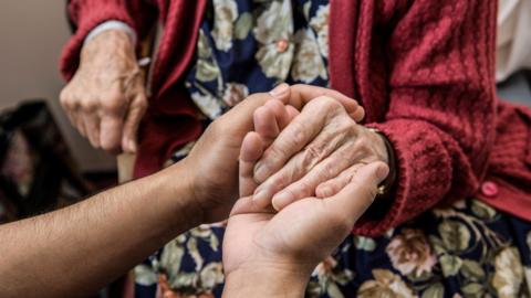 A close-up of two people holding hands. One of the hands belongs to an older person, who wears a floral dress and red cardigan.