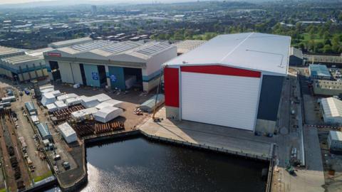 A drone photo of a large red and grey warehouse on a sunny day, with water in front and city buildings trailing off into a tree-lined horizon