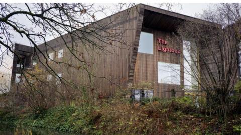 A brown building with a red sign reading "The Lightbox" on it. the building is shielded by a grass verge and trees with no leaves on.