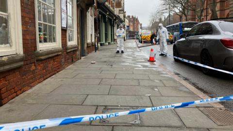 A street in the old quarter of Derby, showing a cordon of police tape, behind which people work in white forensic suits.