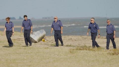 Five people wearing navy shirts and trousers walking in a line along the sand. The sea is behind them and there is a bouquet of yellow sunflowers on the sand.