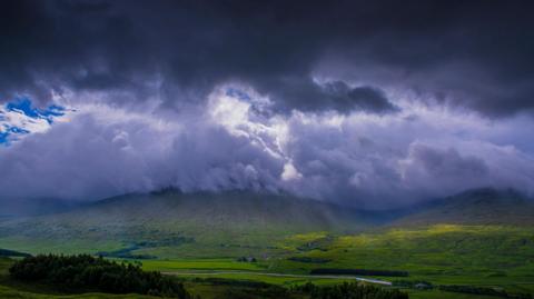 Blue sky peeking through dark ominous clouds over a hilly landscape