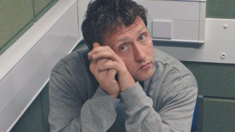 CCTV image of france in police custody. He is in a police interview room with grey brick walls. He is wearing a grey jumper and has curly brown hair. His elbows are resting on the table with his hands up against the left side of his face, He is looking directly at the camera.