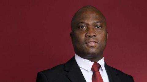 Dominique Désiré Erenon stands in front of a dark red background. He wears a white shirt, red tie, black suit and faces the camera.