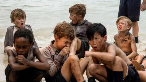 Six boys sit or crouch on a sandy beach near the water, their clothes and skin dirty, some appearing tired.