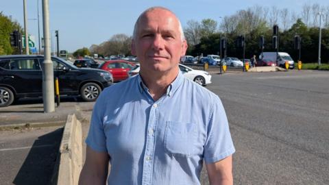Councillor Sean Rhodes wearing a short-sleeved pale blue shirt. He has a bald head and is clean shaven, standing beside a road with lots of cars queuing. It is a sunny day and he is smiling at the camera.