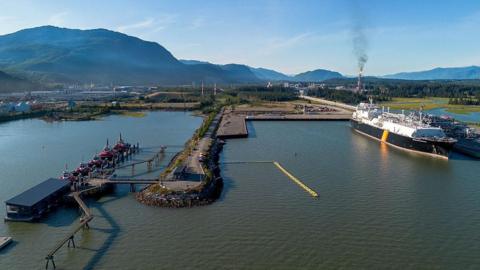 An image showing an LNG carrier loaded at the LNG Canada docks in Kitimat, British Columbia. There is port infrastructure visible, a large ship and LNG flares in the background emitting from the facilitiy. It is set on the water, with a picturesque backdrop of small mountains and pristine, green landscape.