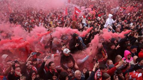 A large crowd of Liverpool fans are holding smoke bombs in a crowd as they watch the parade bus pass