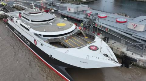 The fastcraft Manannan Steam Packet Company vessel in white and black, is docked alongside a long building with the Manx triskelion displayed on the left, right and centre of the building. People in high vis jackets can been seen on an elevated walkway connecting the ship to the land.